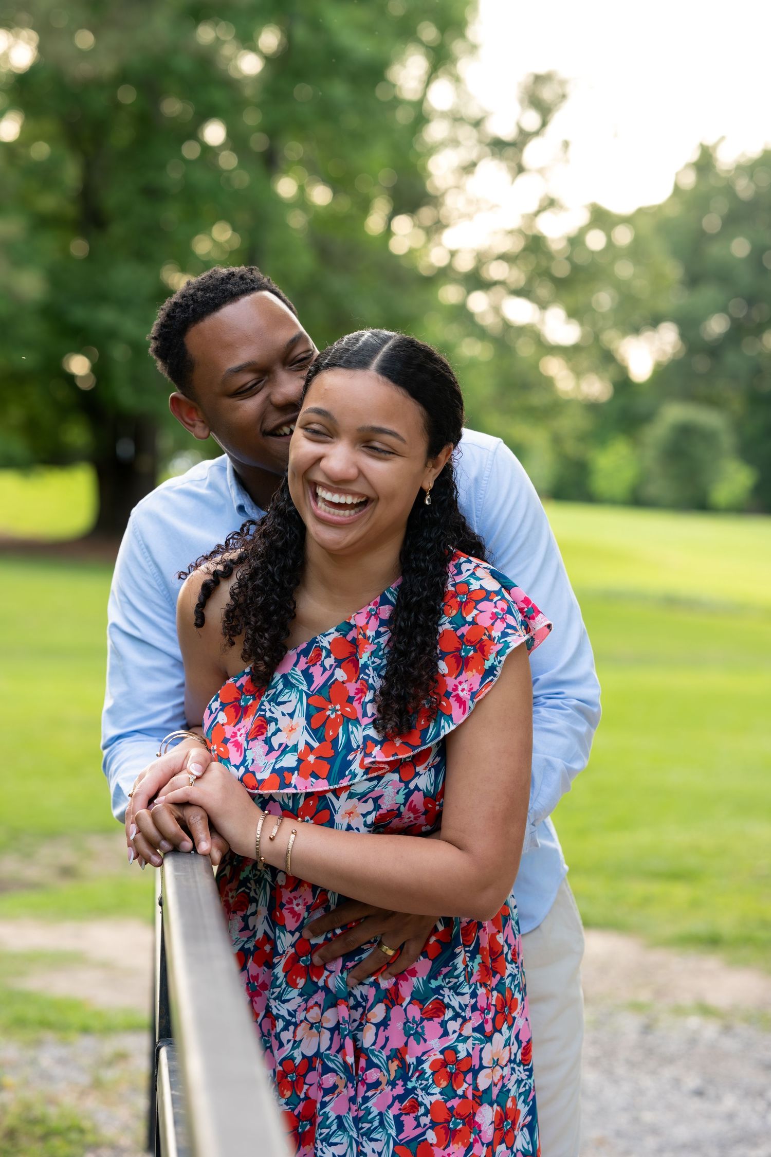 A joyful couple embraces outdoors, woman in floral dress laughing as man kisses her cheek in a lush green park.