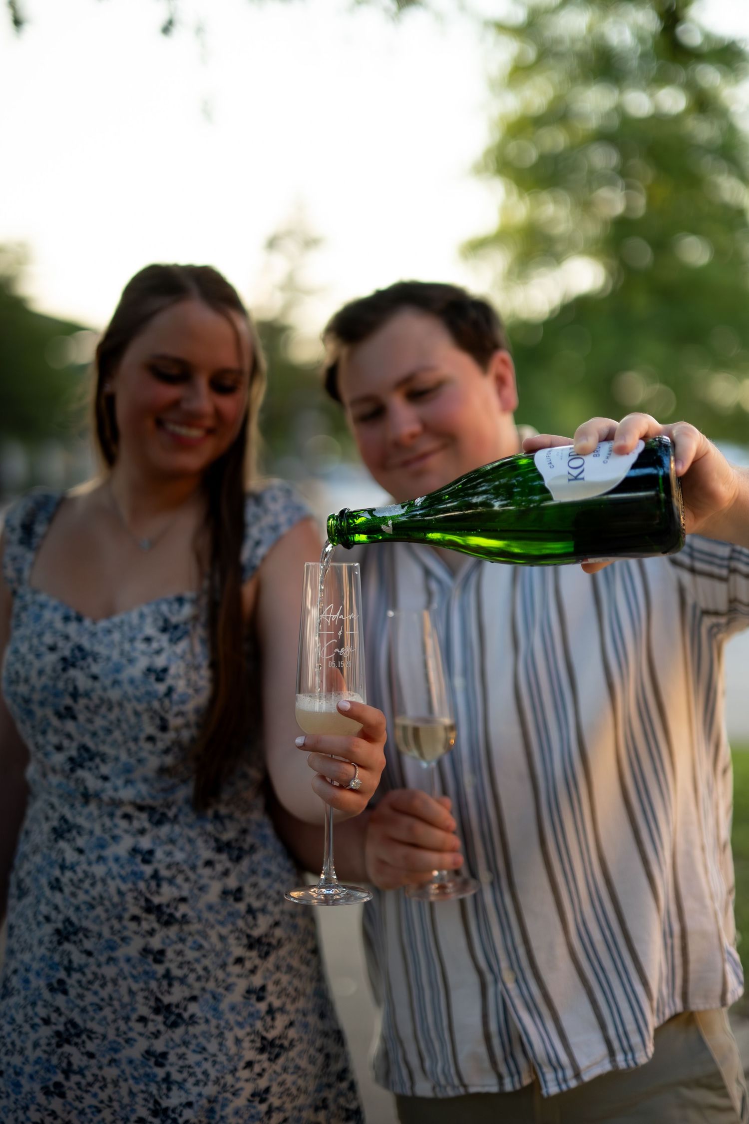 Couple celebrating outdoors, man pouring champagne into flute glasses at sunset with green trees in background.