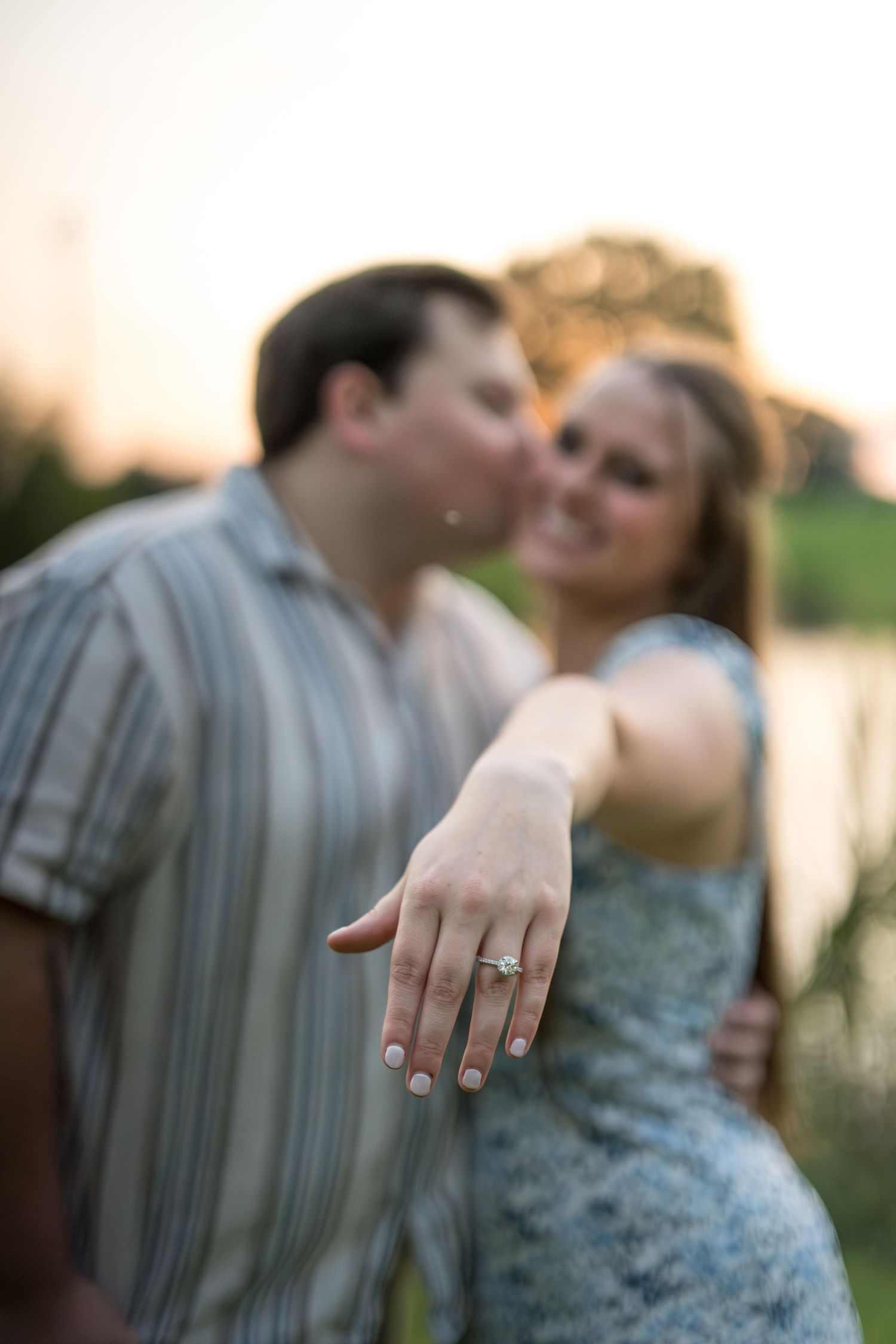 Engaged couple kissing while woman shows off diamond engagement ring at sunset.