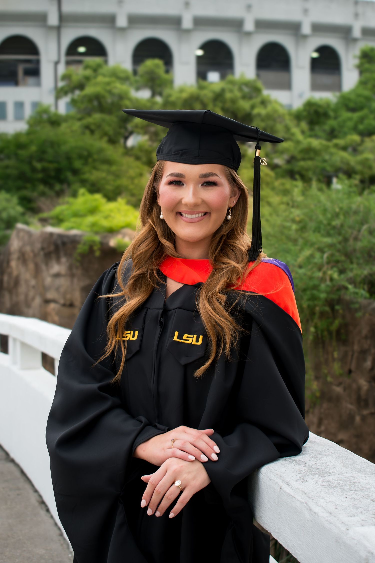 Smiling woman in LSU black graduation cap and gown with red and gold hood posing outdoors near a stadium.