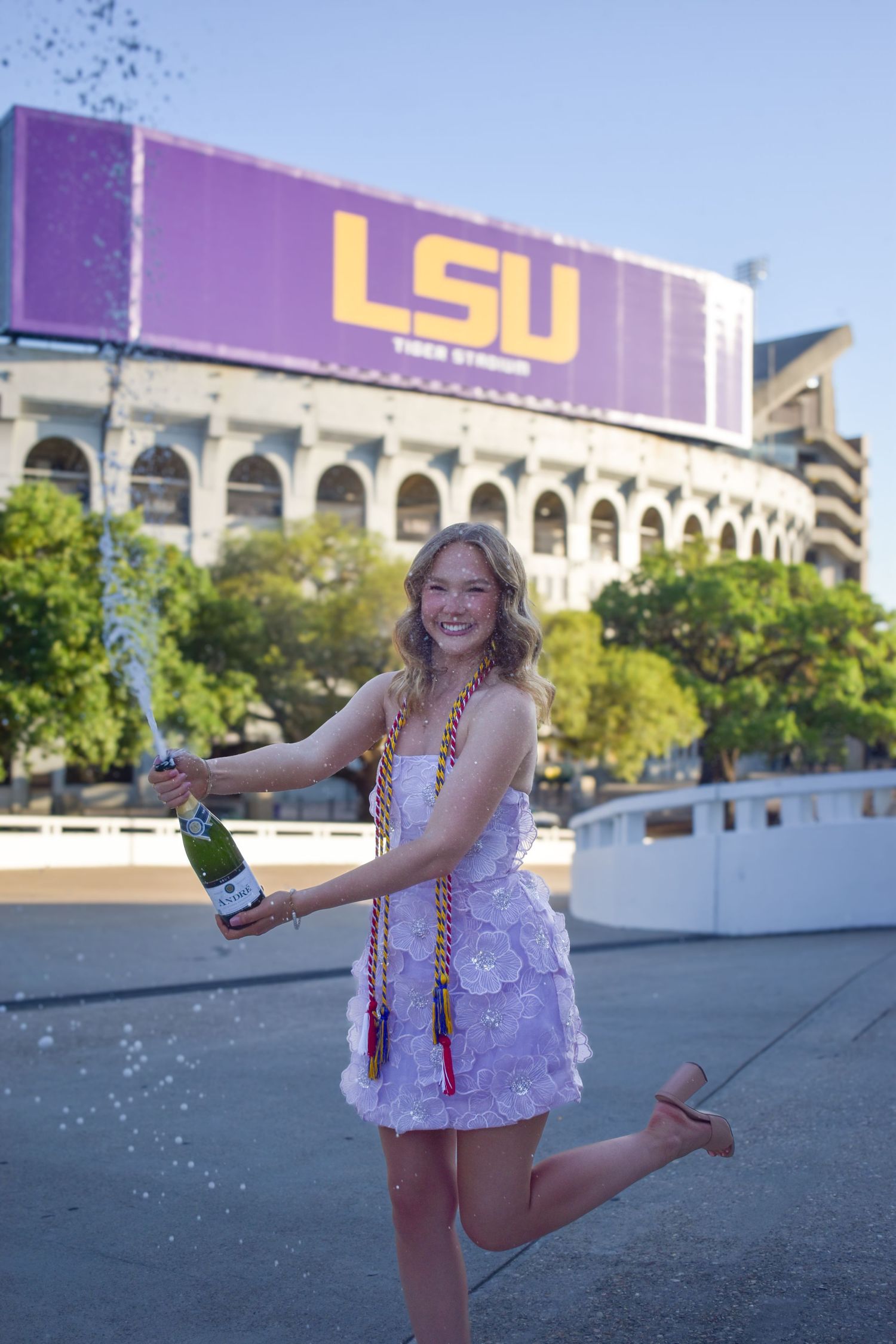 Smiling graduate pops champagne bottle in front of LSU Tiger Stadium sign.