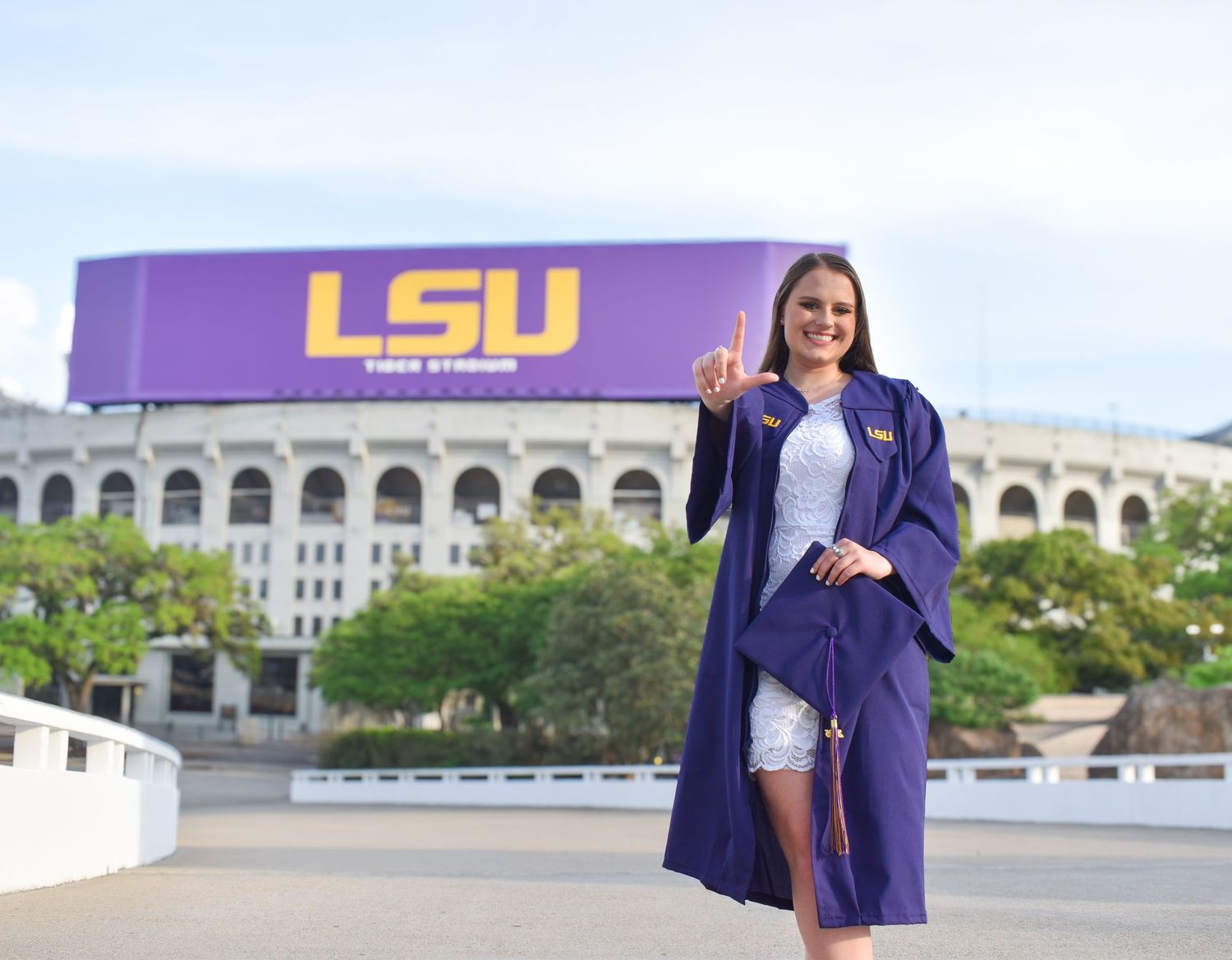 LSU graduate in purple gown poses in front of Tiger Stadium holding up number one finger.