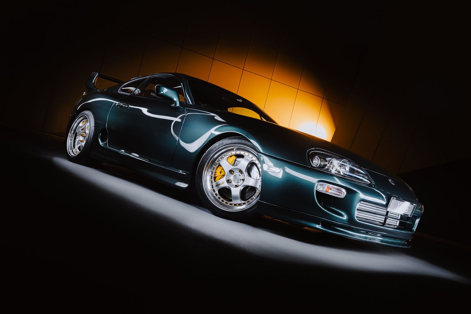 Dark green sports car with chrome wheels displayed in dramatic studio lighting against a golden background.
