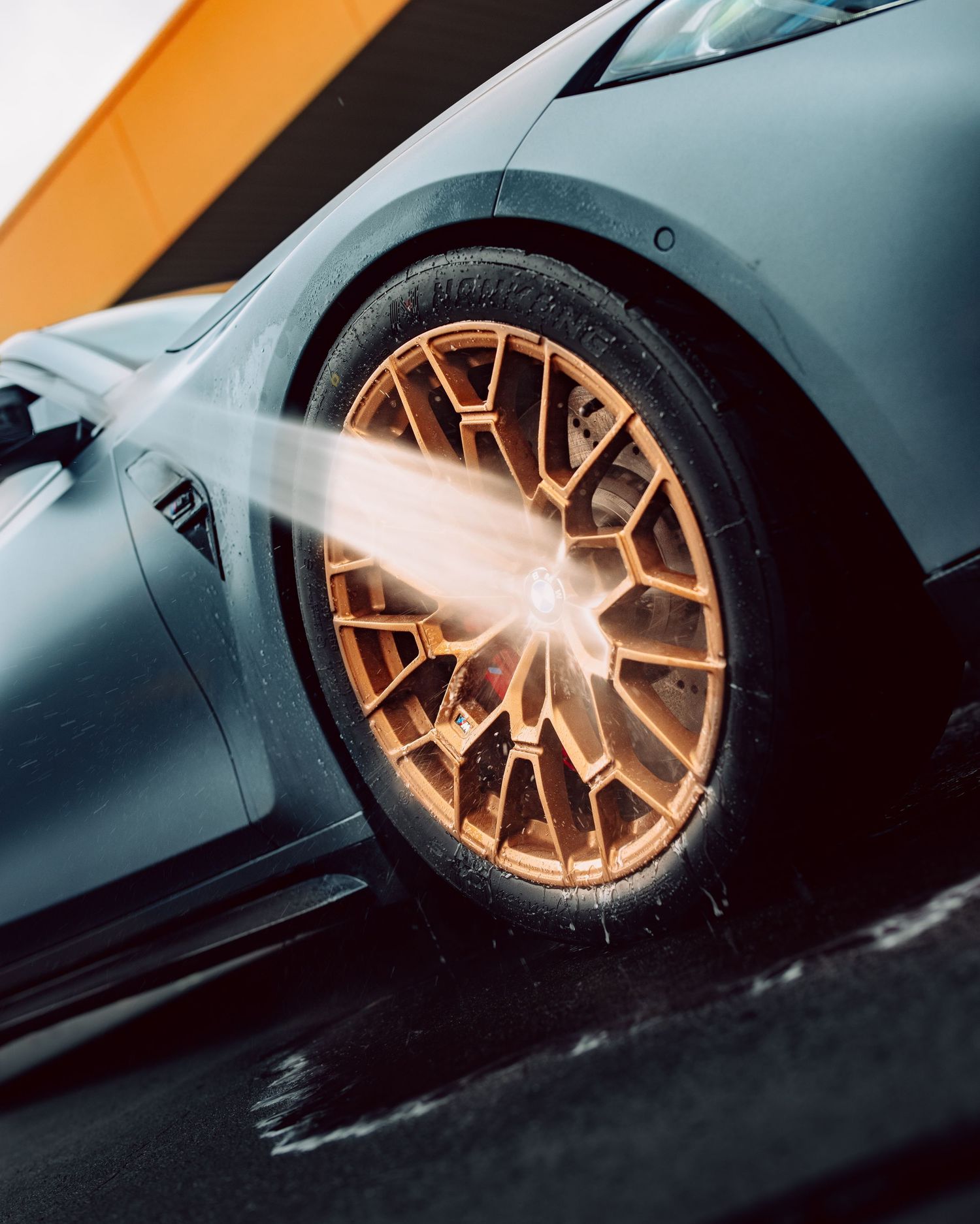 A bronze multi-spoke alloy wheel being pressure washed on a matte gray sports car, with water spray glistening.