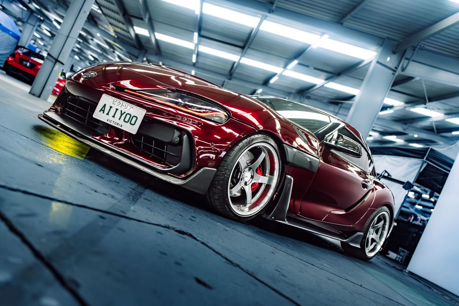 A red modified sports car with custom wide body kit and white wheels parked in a garage showroom.