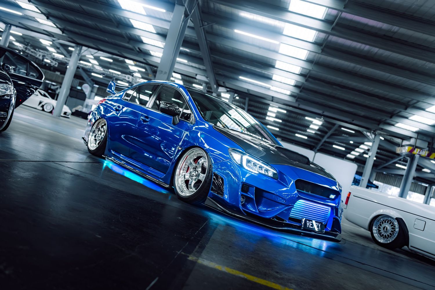 A heavily modified blue Subaru WRX STI with neon underglow lighting sits low on white wheels in a parking garage.