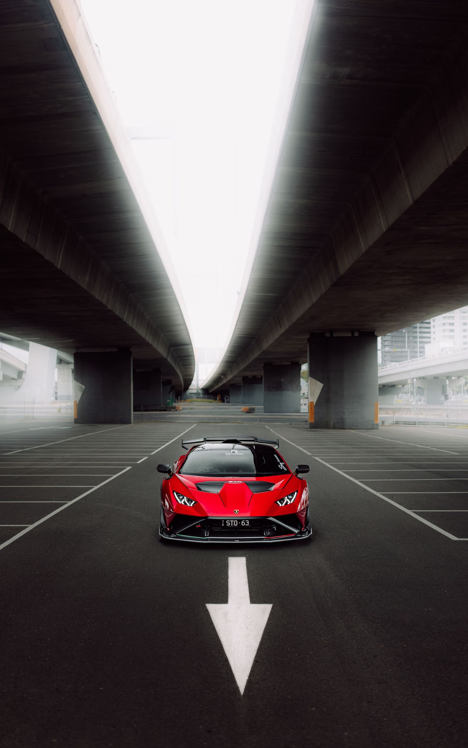A red Lamborghini Huracan supercar facing forward under concrete highway overpasses with an arrow below.