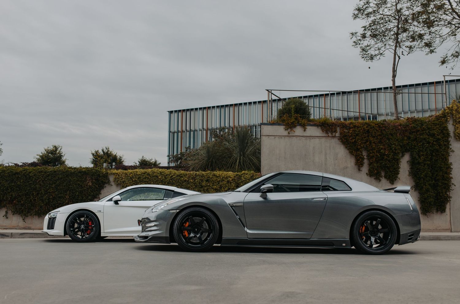 Two sports cars, a white Audi R8 and gray Nissan GT-R, parked side by side in a lot with modern building.