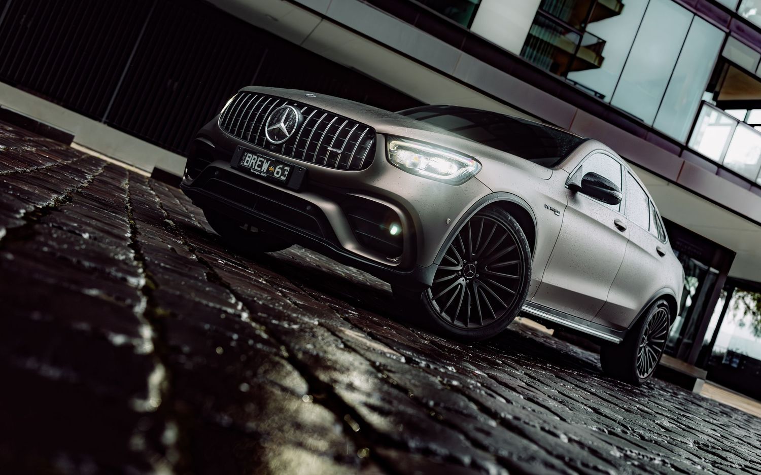 Matte gray Mercedes-AMG SUV with black alloy wheels parked on wet cobblestones in an urban setting.