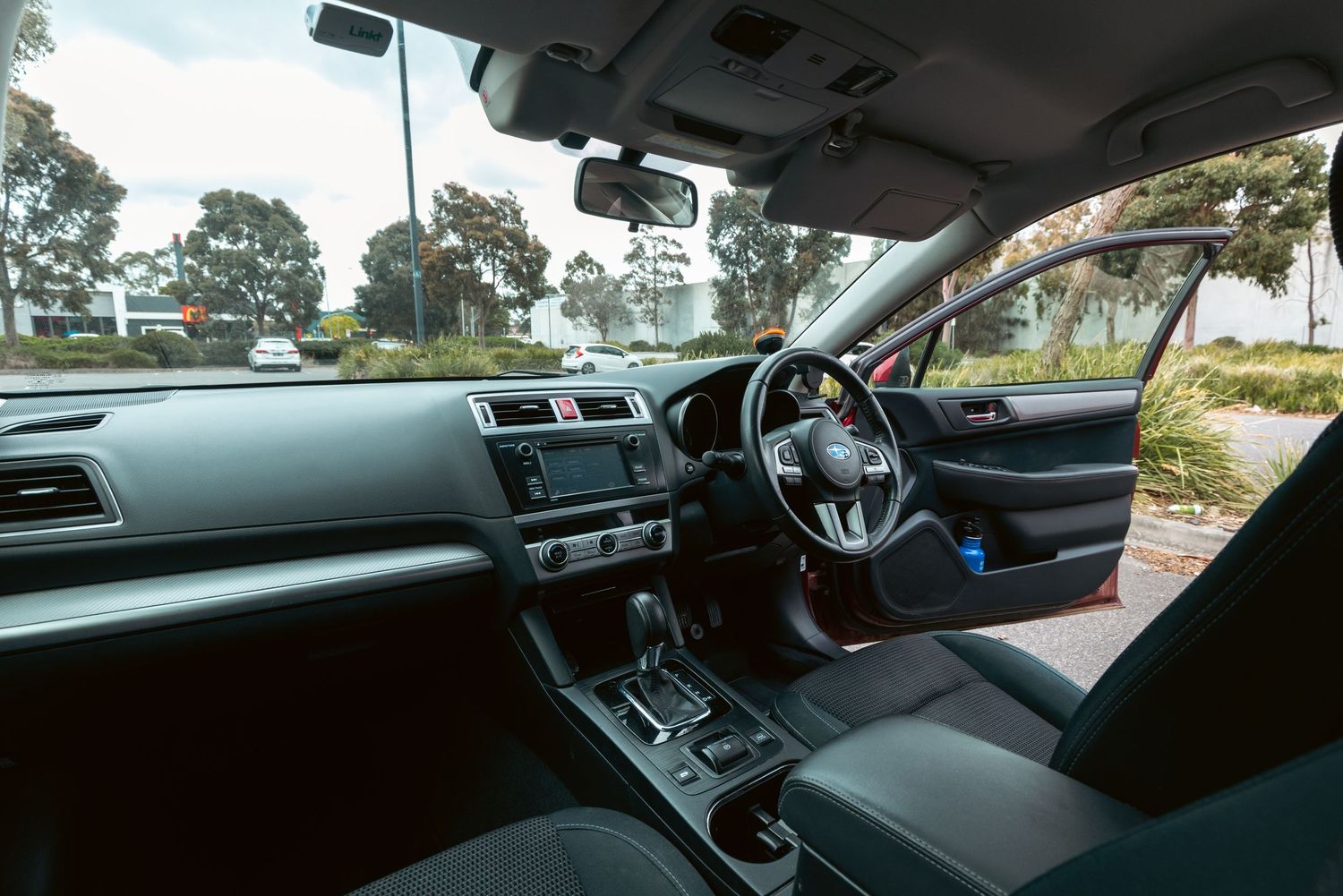 Interior view of a right-hand drive Subaru car showing dashboard, steering wheel, and gear shift with trees visible outside.