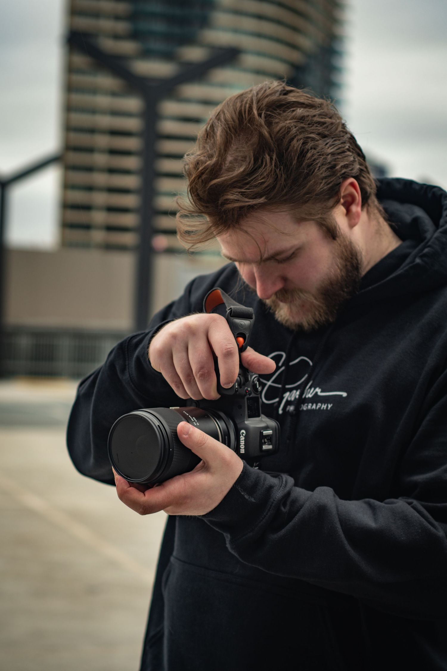A bearded young male photographer adjusts a Canon camera lens outdoors with a city skyline behind.