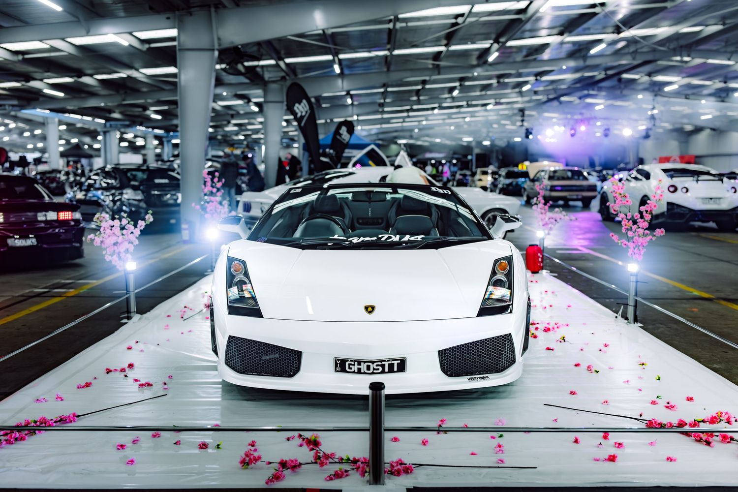 White Lamborghini Gallardo wedding car decorated with pink rose petals in an indoor parking venue with festive lights.