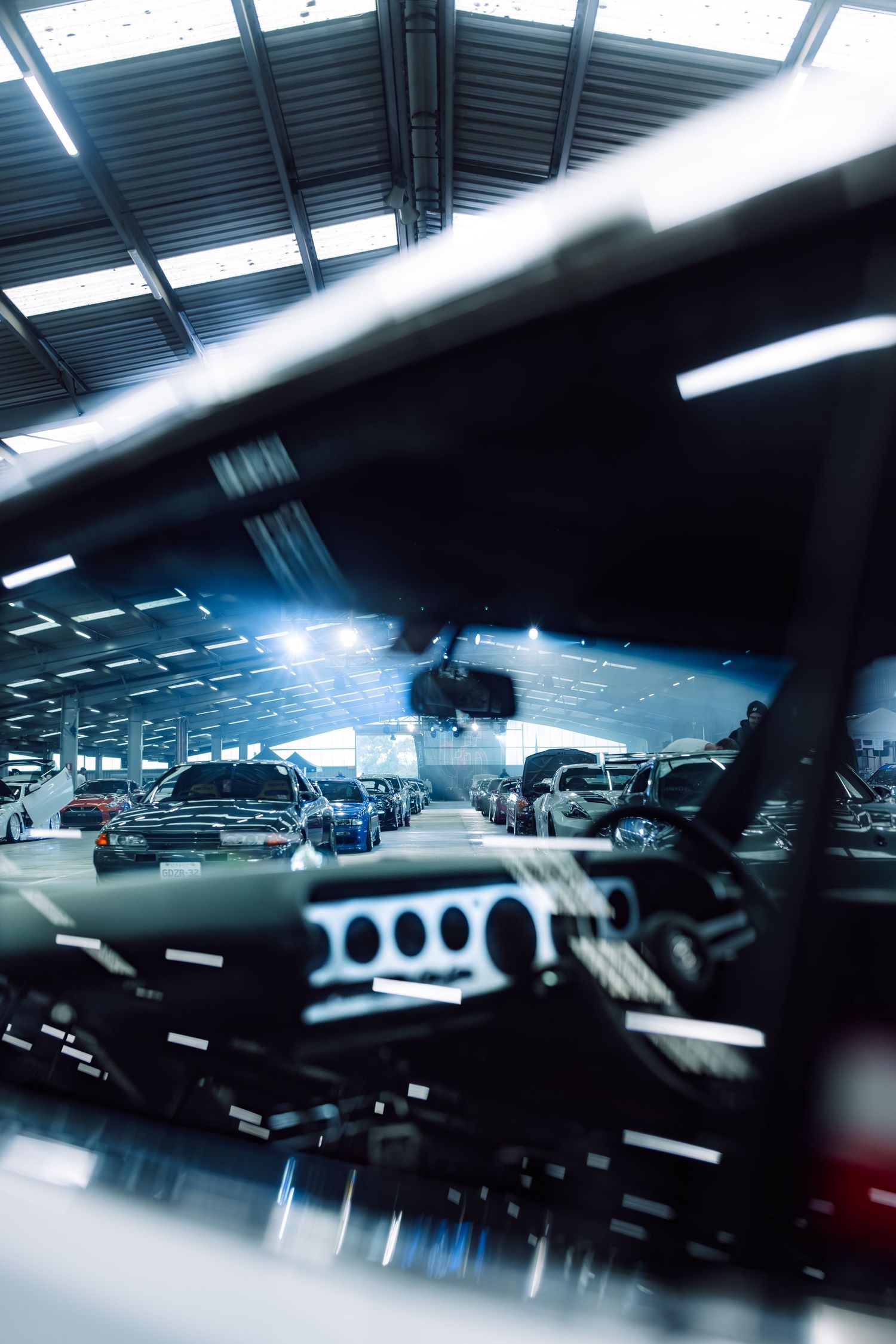 Interior view through a car windshield inside a large automotive showroom with multiple vehicles and bright overhead lighting.