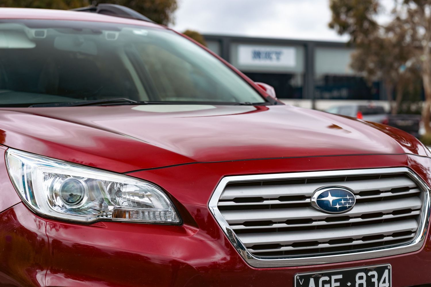 Red Subaru Outback SUV close-up showing chrome grille, headlight, and blue star logo badge with license plate AGE-834.