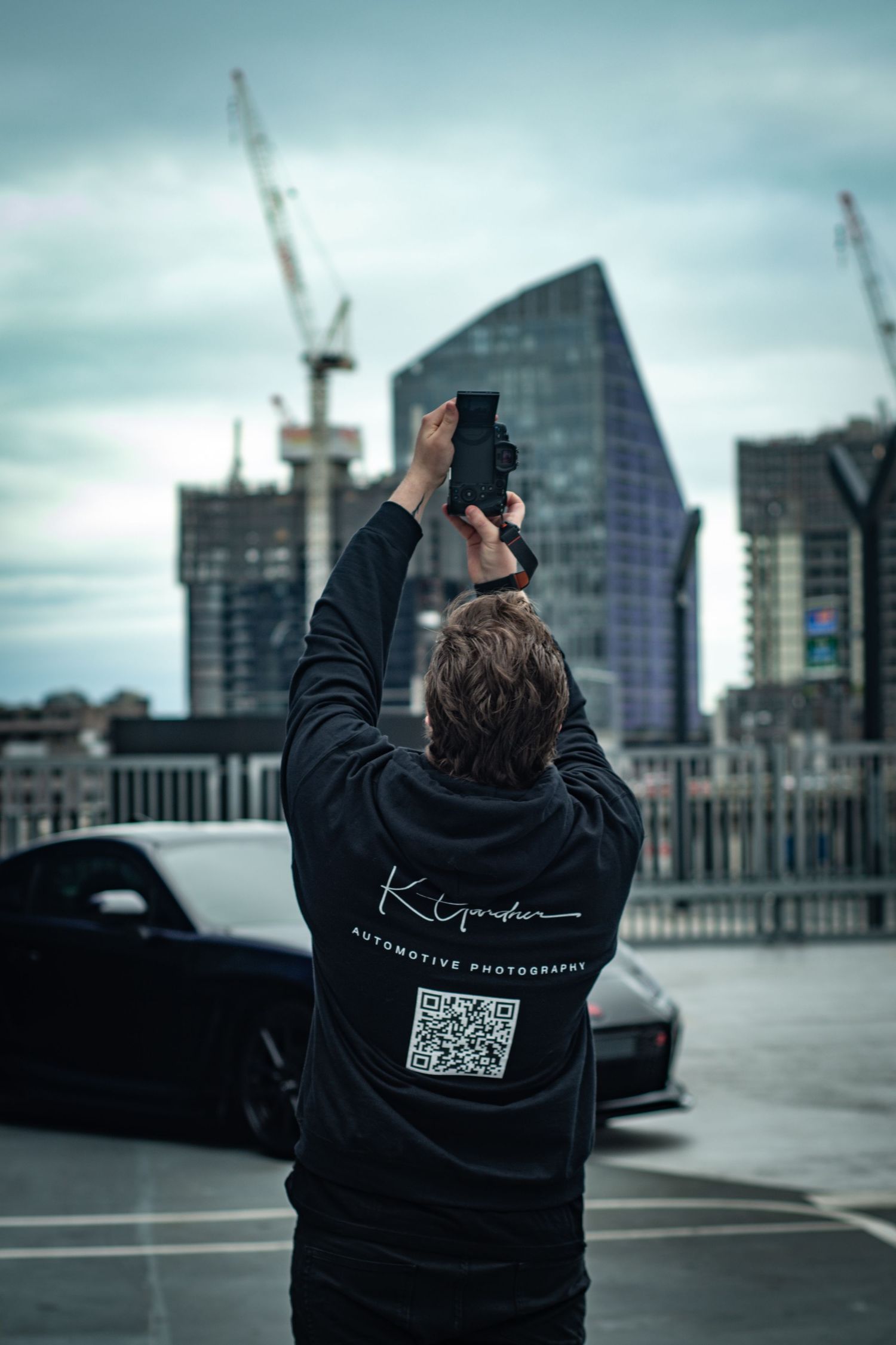 Photographer in black hoodie with QR code holds camera overhead on rooftop with city skyline and cranes in background.