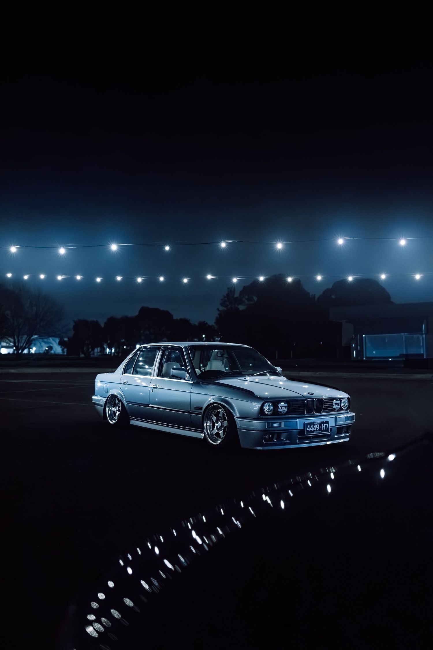 Silver BMW E30 sedan parked at night under string lights, showcasing a classic modified stance on dark pavement.