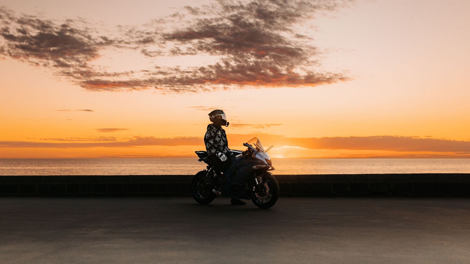 Motorcyclist silhouetted against a stunning ocean sunset on a coastal road.
