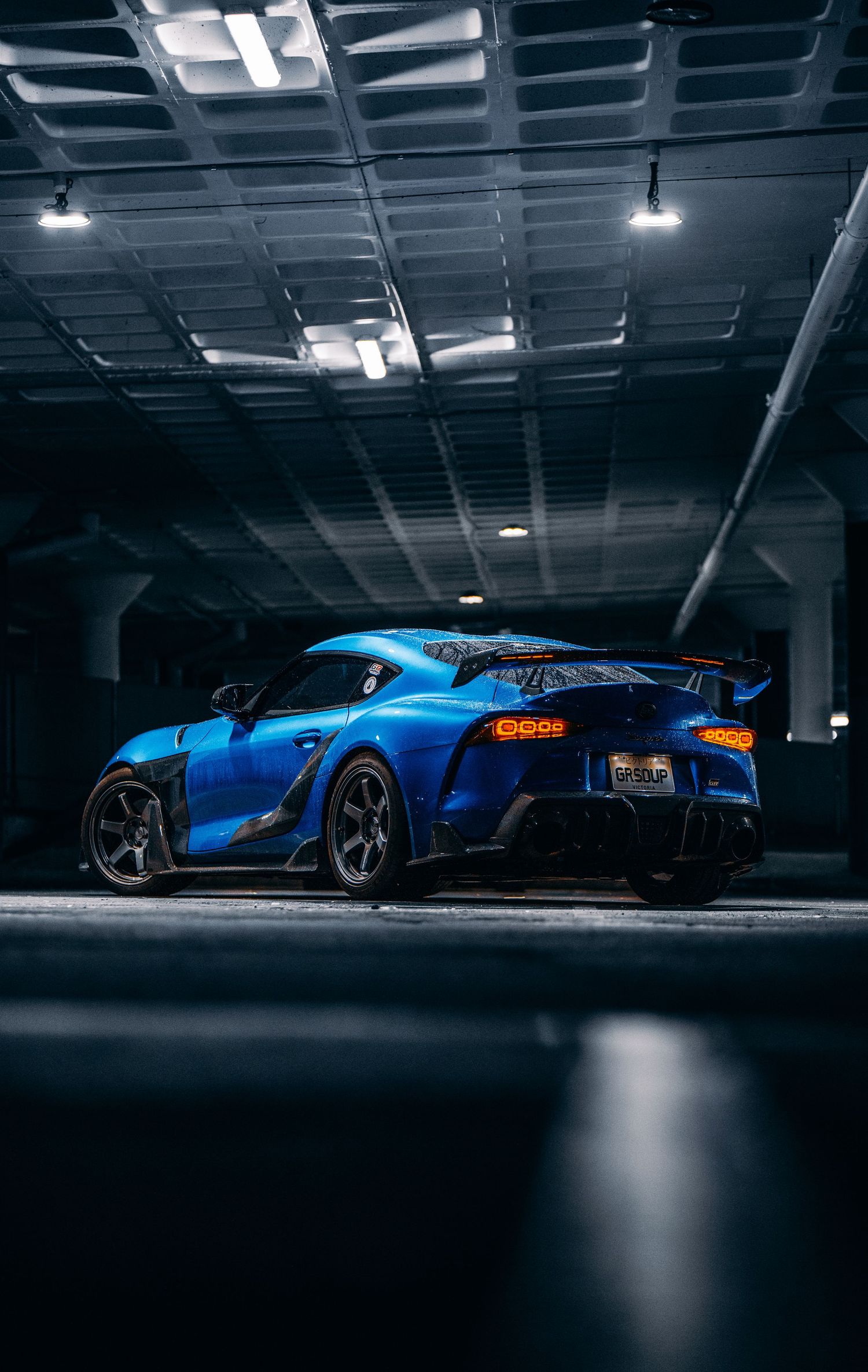 A blue Toyota Supra with a large rear spoiler parked in a dimly lit underground parking garage.