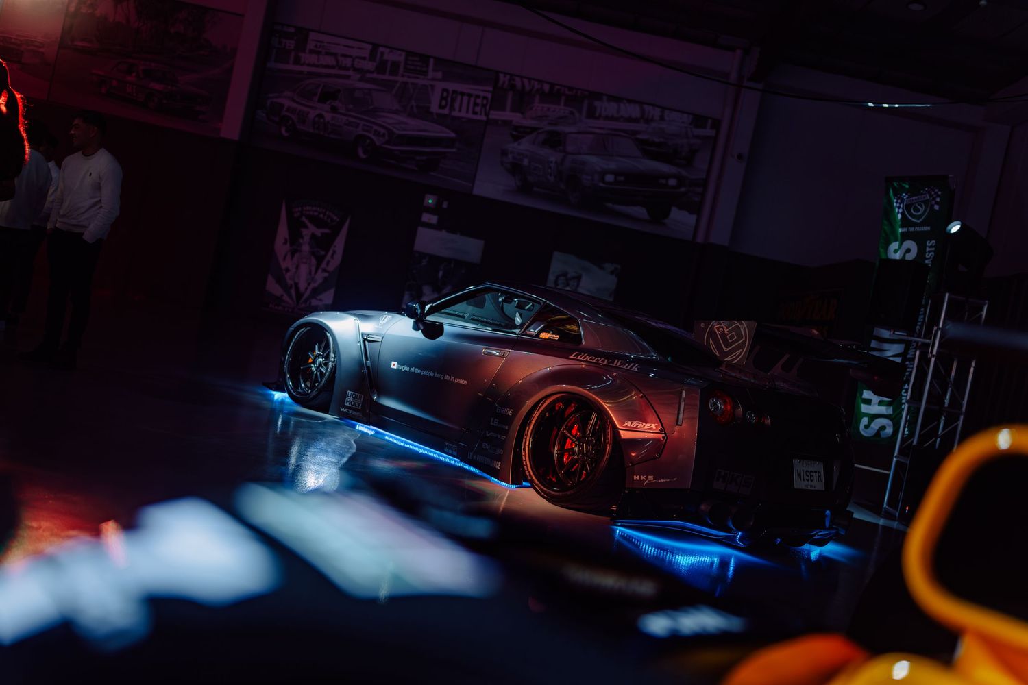A sleek dark sports car displayed under dramatic blue neon lighting at an indoor automotive show.