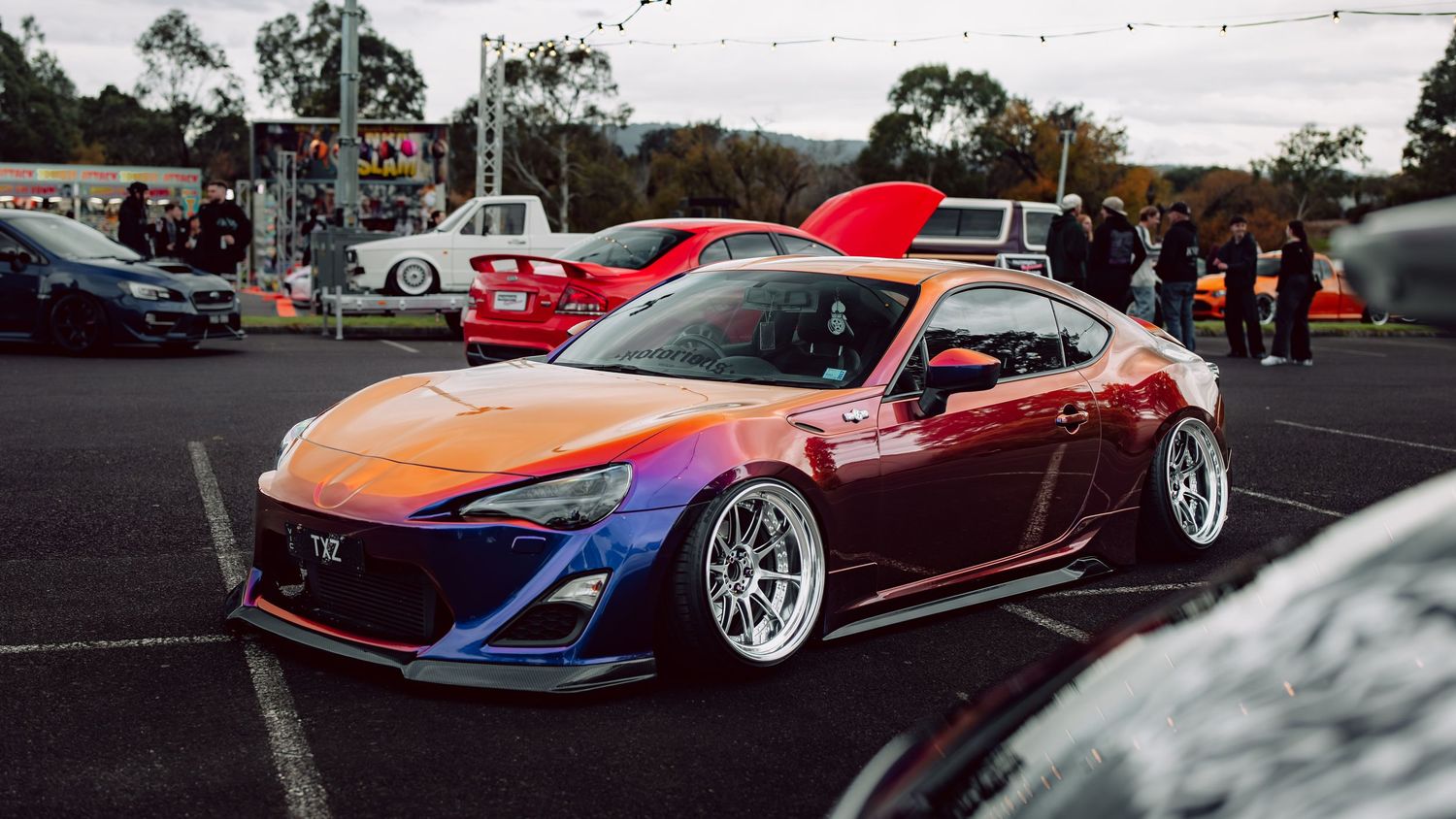 A heavily modified Toyota 86 with iridescent wrap and wide body kit sits low on BBS wheels at a car meet.