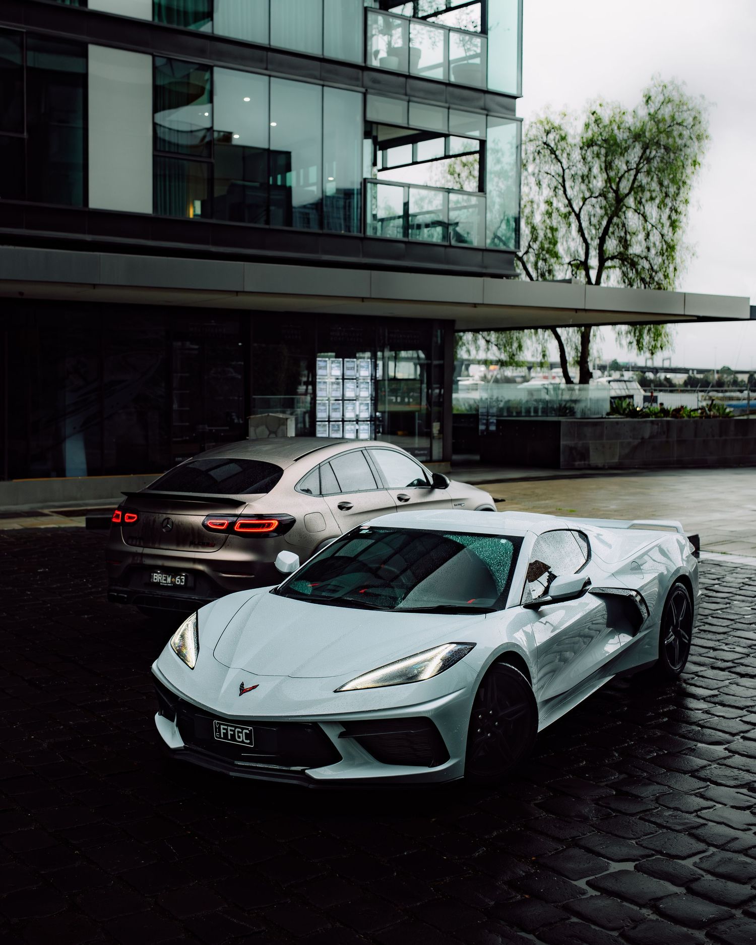 White Chevrolet Corvette C8 parked on cobblestone street beside modern glass building with luxury sedan in background.