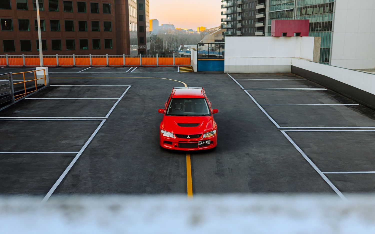 Red Mitsubishi Lancer Evolution parked on rooftop parking lot at sunset with city buildings in background.