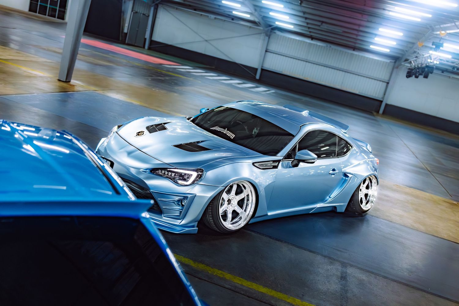 Light blue modified Subaru BRZ with wide body kit and lowered stance parked in a garage.