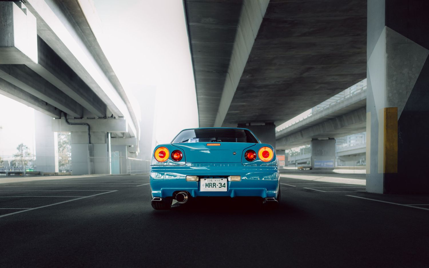 Blue Nissan Skyline GT-R R34 rear view under concrete overpass with glowing red taillights on empty road.