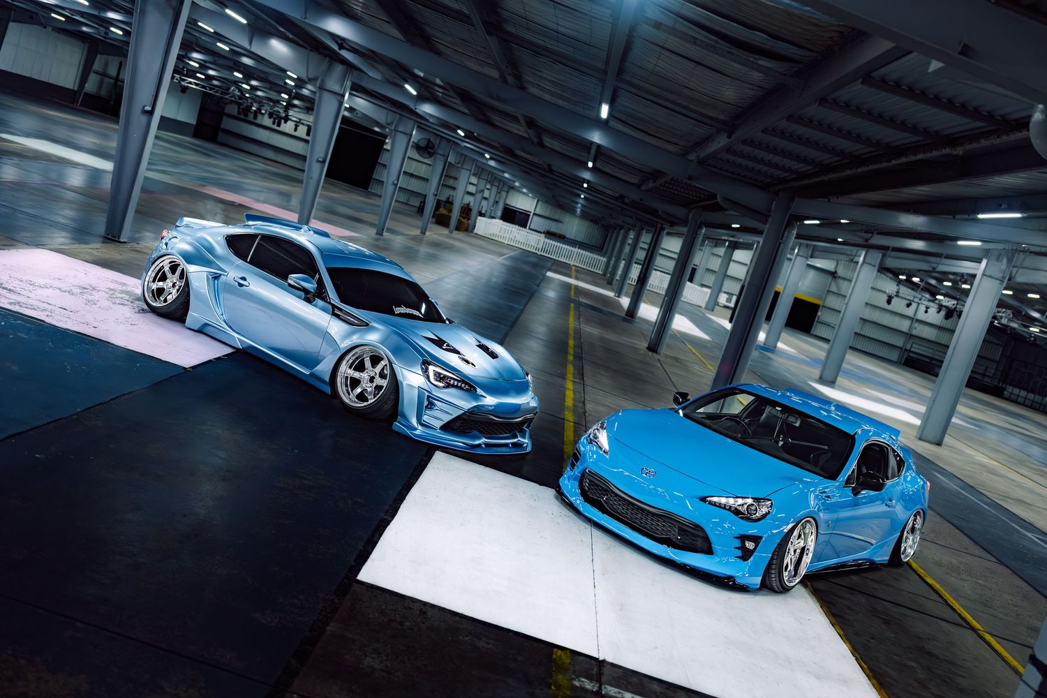 Two custom baby blue Toyota 86 sports cars parked in a modern parking garage under dramatic lighting.