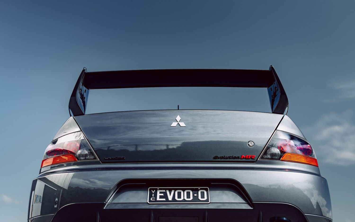 Rear view of a Mitsubishi Lancer Evolution with a large spoiler and EVO-0 license plate against a blue sky.