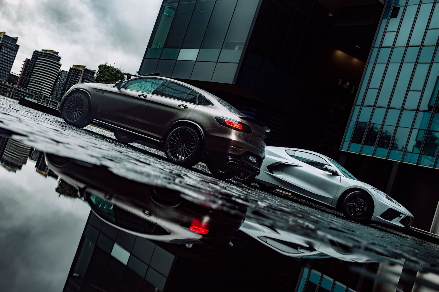 Two sleek luxury sports cars reflect on wet pavement near modern glass buildings under dramatic cloudy skies.