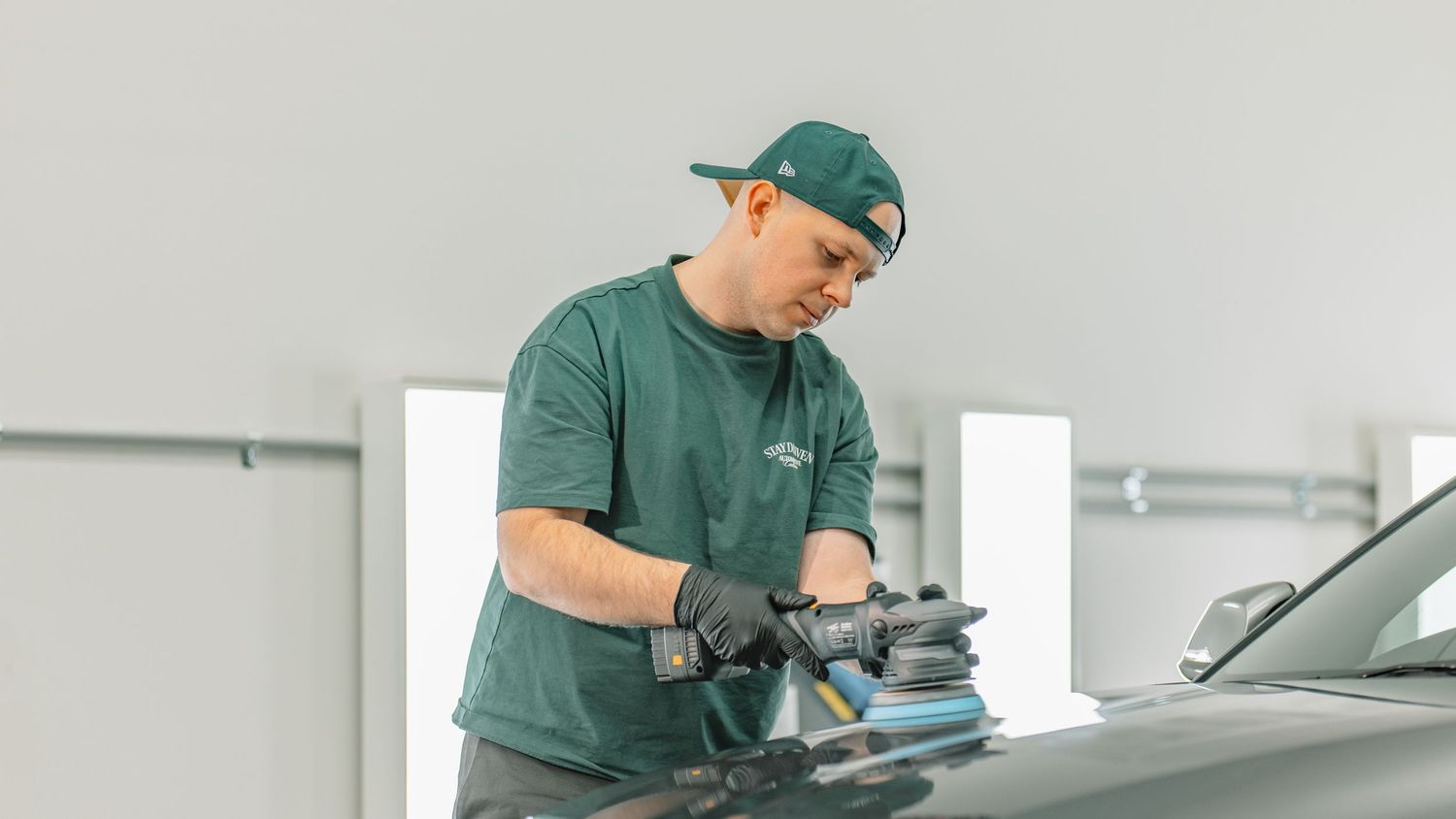 Auto detailer in green shirt polishing a car hood with a dual-action polisher.