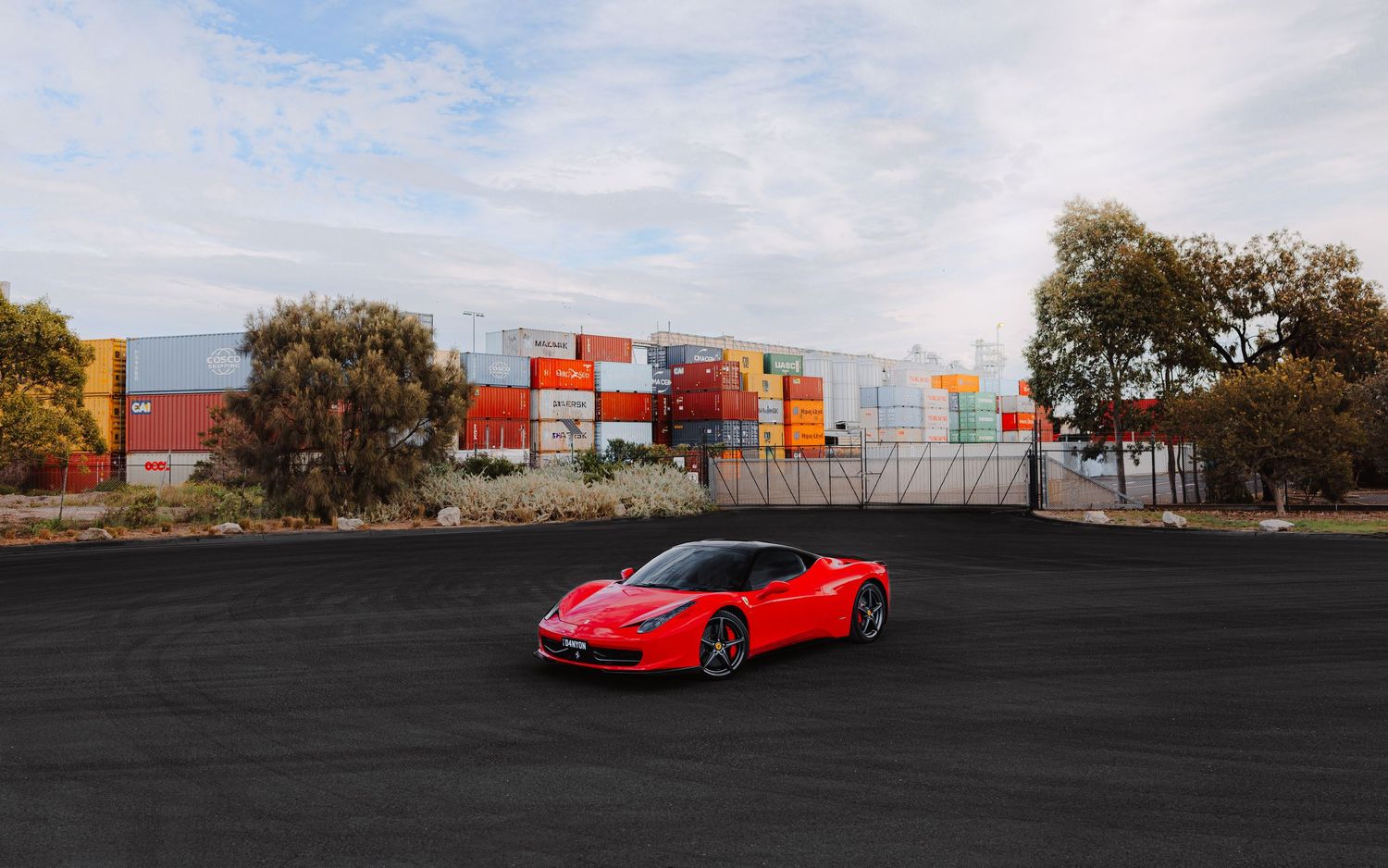 Red Ferrari 458 parked on dark asphalt with colorful shipping containers and trees in the background under a cloudy sky.