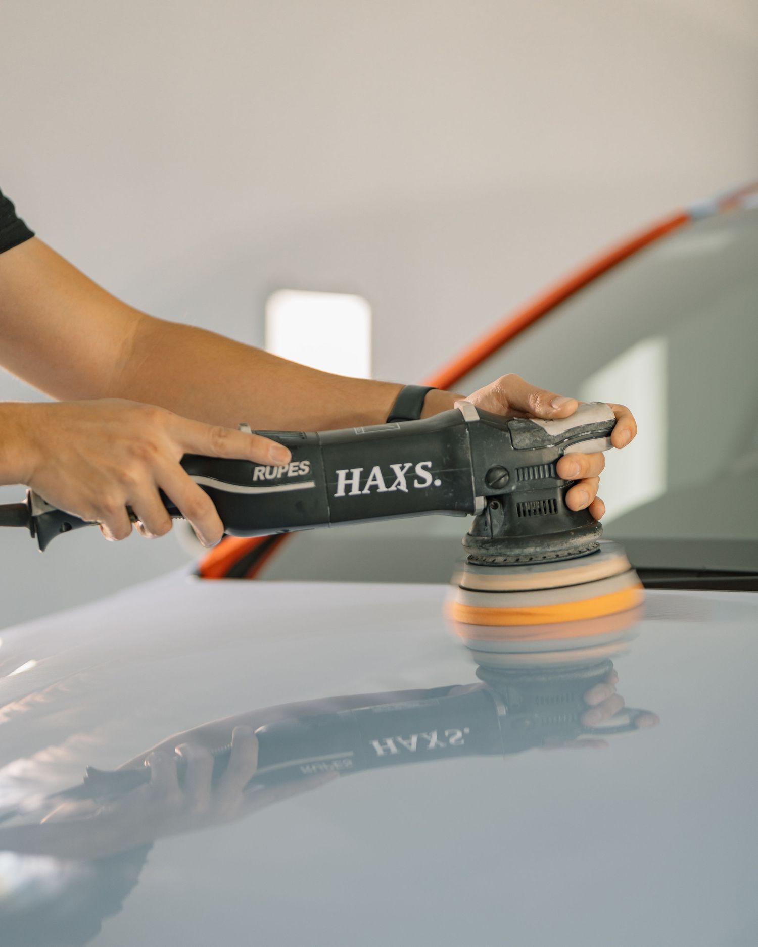 Close-up of hands using a RUPES orbital polisher with an orange pad on a blue car surface.