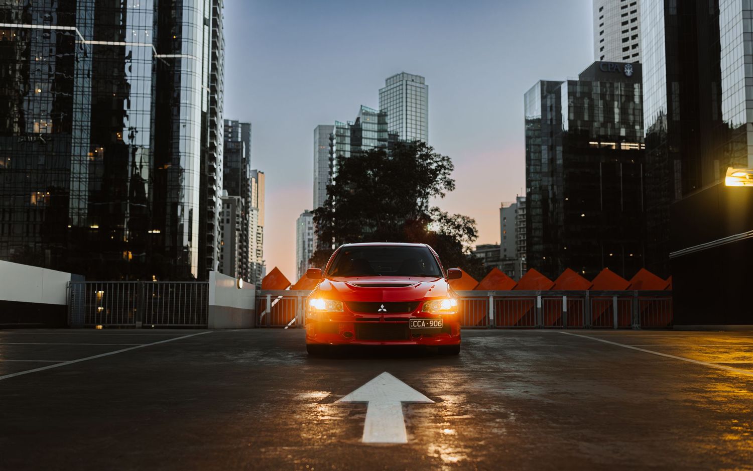 A red Mitsubishi Lancer parked on a city street with glowing headlights, skyscrapers, and a white arrow painted on the pavement.
