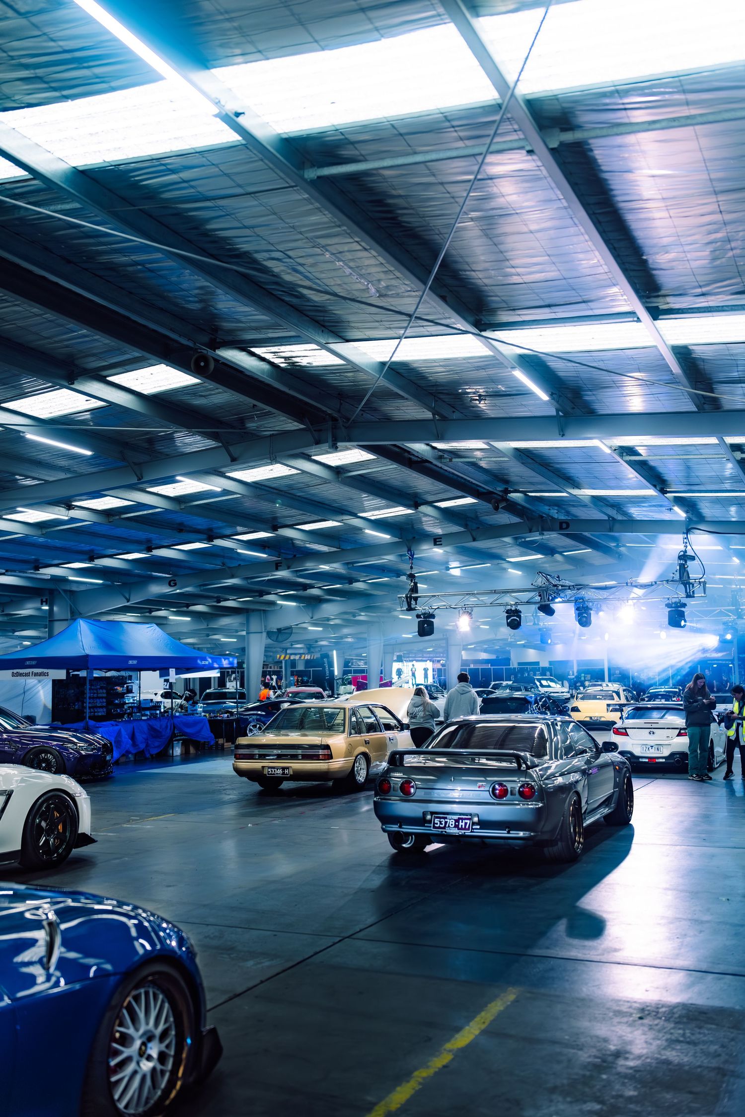 Indoor car show featuring modified vehicles including a Nissan Skyline, classic cars, and sports cars under blue-tinted warehouse lighting.