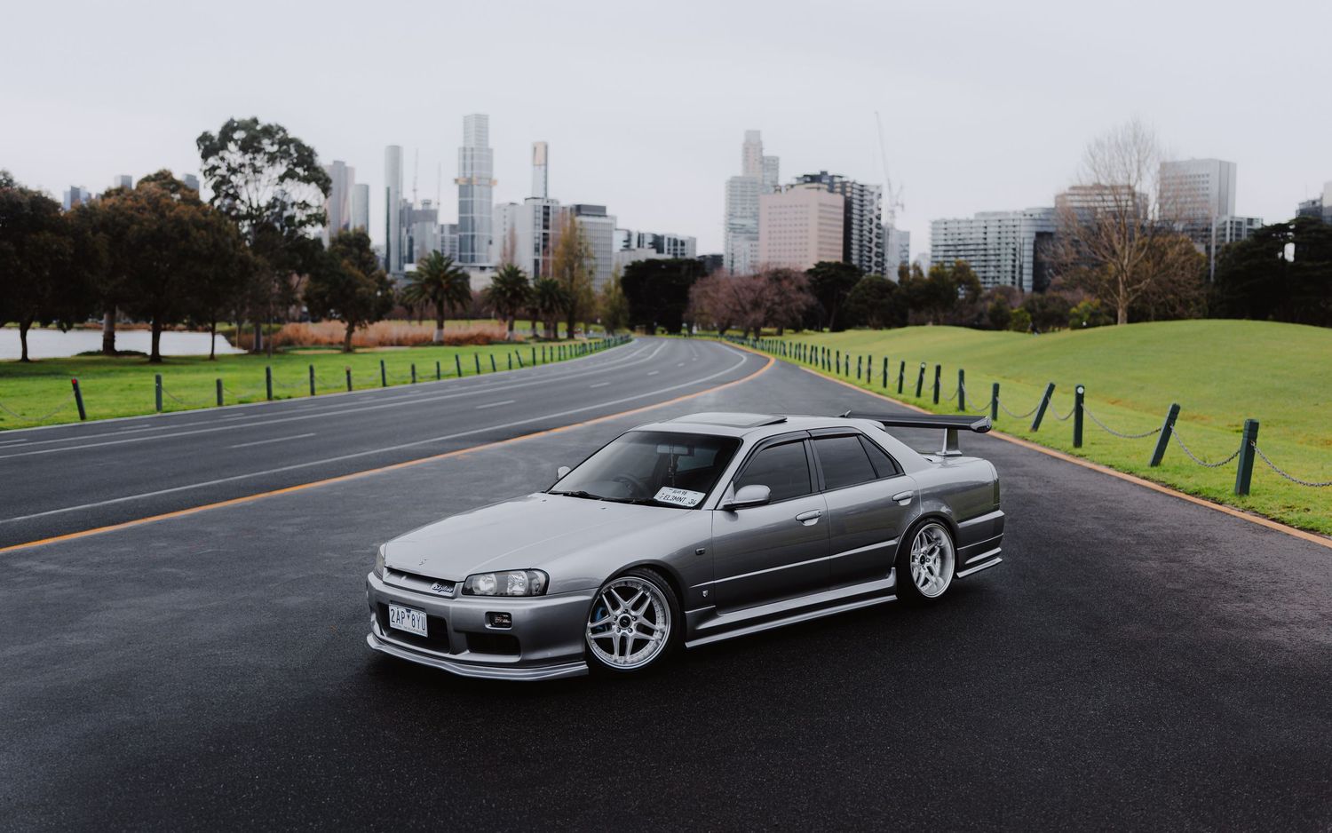 Silver Nissan Skyline R34 parked on a wet road with lush green grass and city skyline in the background.