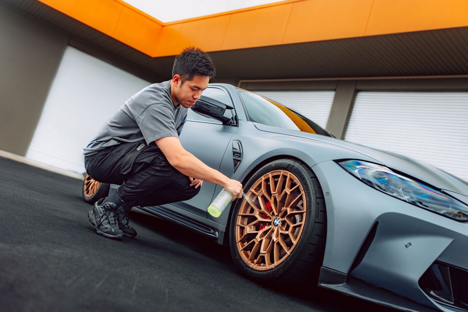 Man detailing a matte gray sports car with bronze alloy wheels in a modern garage setting.