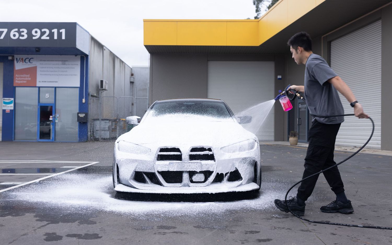 A man pressure washing a white BMW sports car with foam soap outside an auto detailing shop.