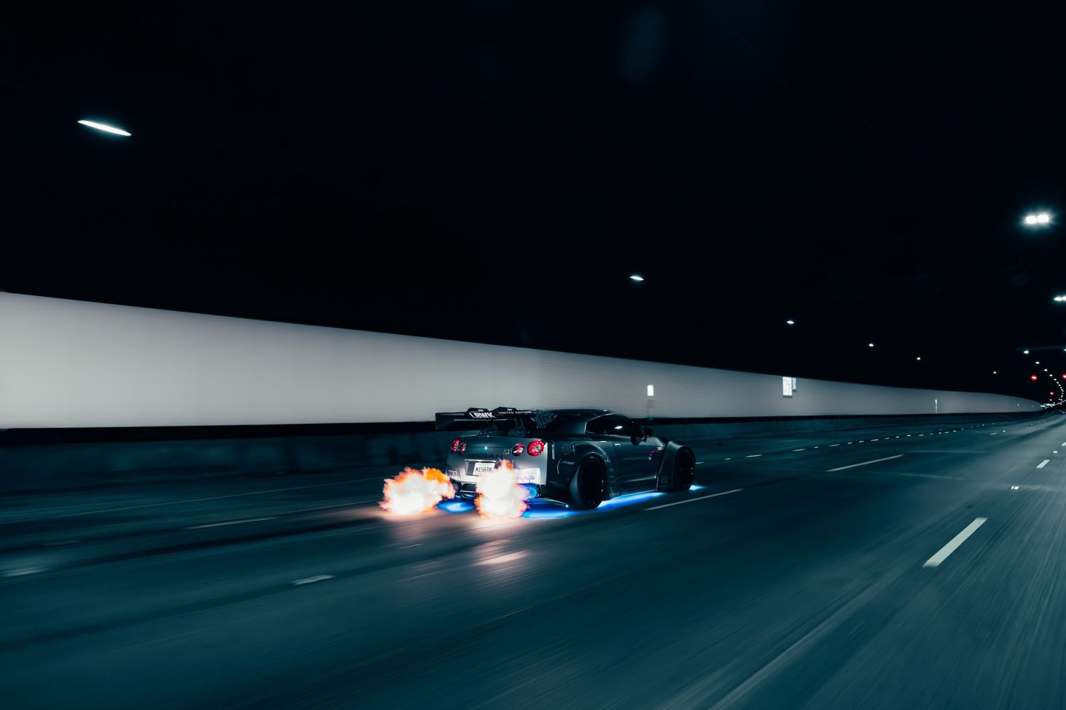 A sports car speeds through a dark tunnel at night, flames shooting from its exhaust pipes on a wet road.