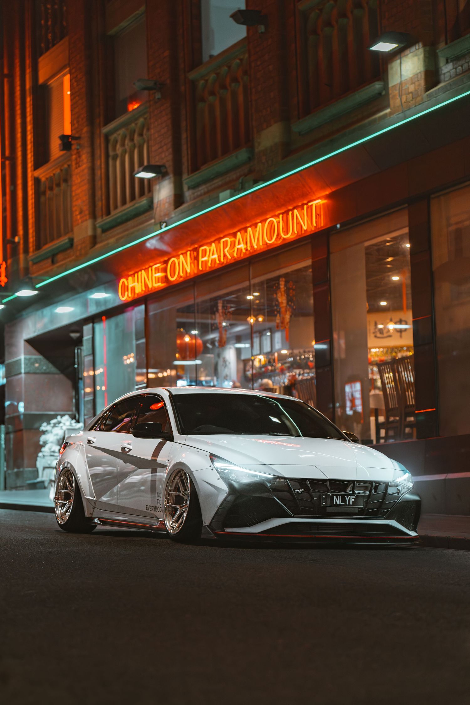 A modified white Hyundai Elantra N stance car parked outside a neon-lit restaurant at night.