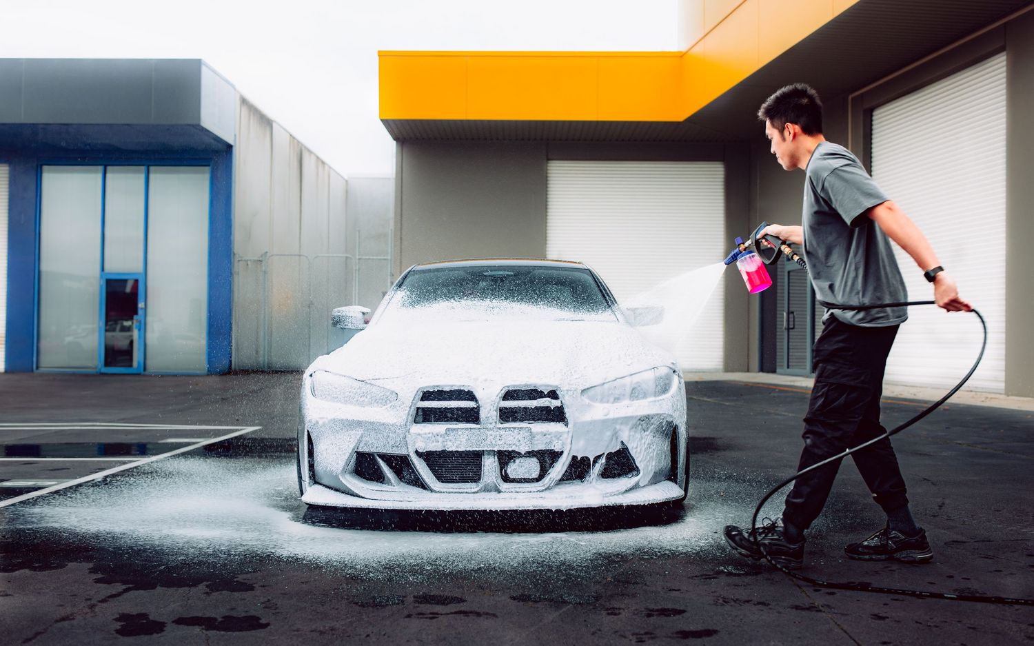 Man pressure washing a white BMW sports car with pink foam cannon at an outdoor car wash facility.
