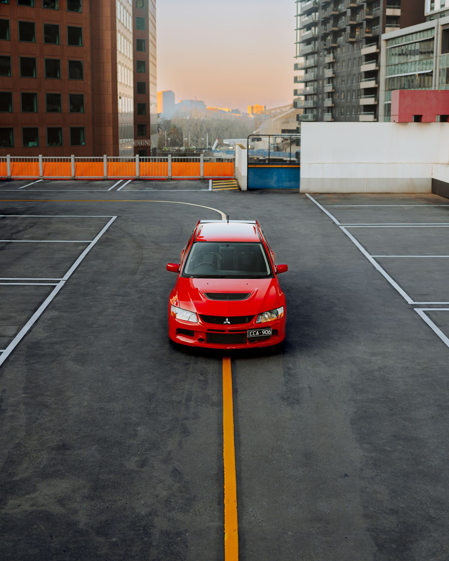 Red Mitsubishi Lancer Evolution parked on rooftop parking lot at sunset with city buildings in background.