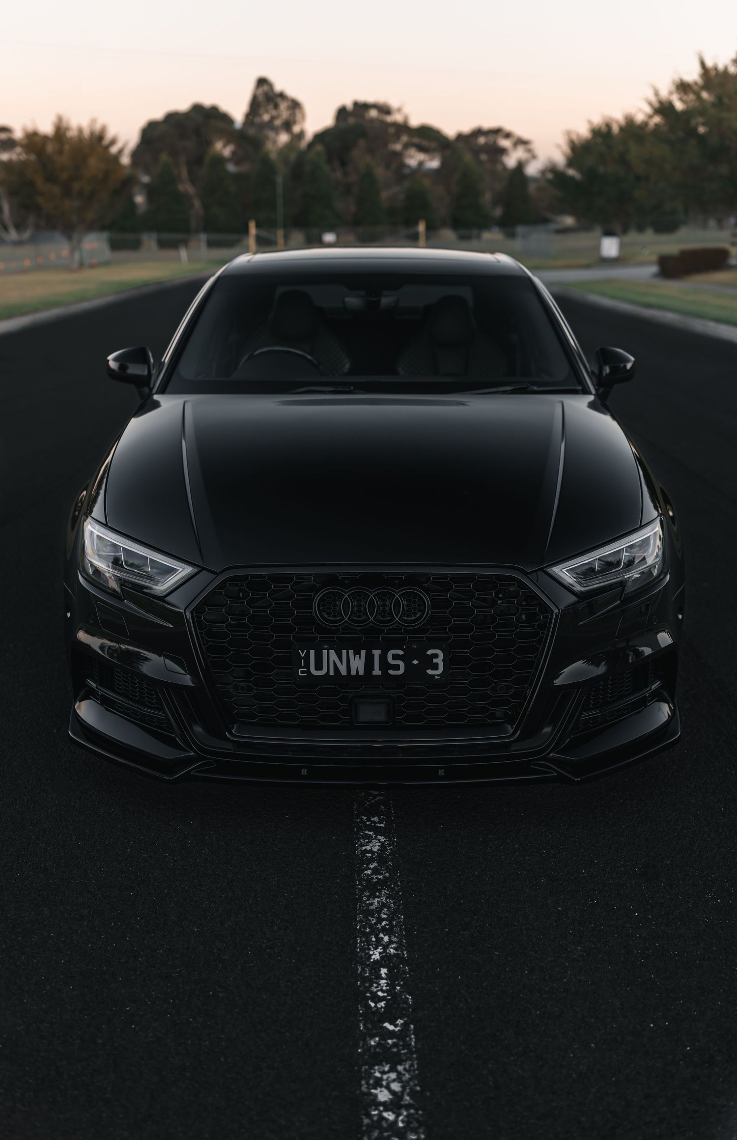 Sleek black Audi RS3 facing forward on wet road at dusk with dramatic lighting.