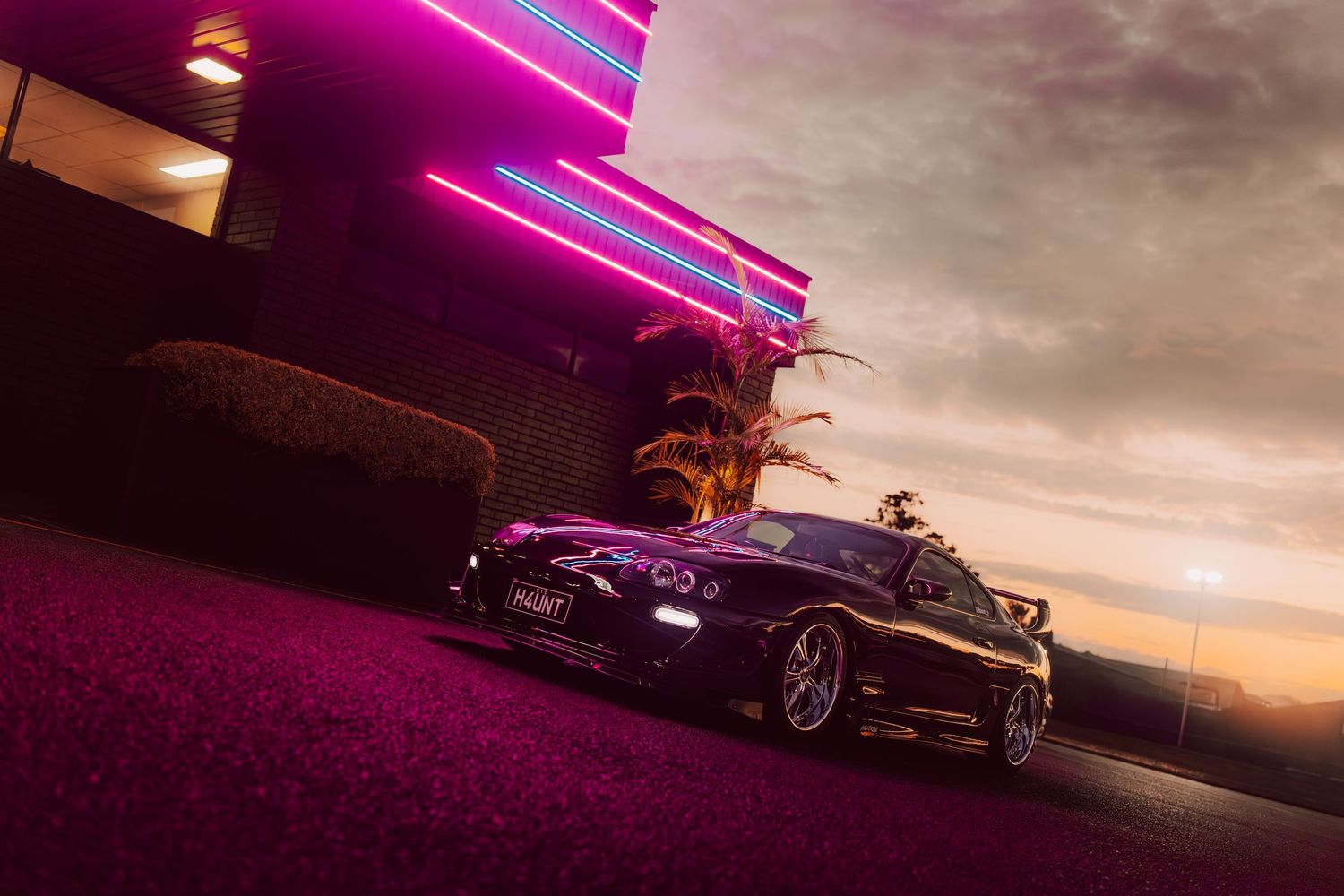 Black sports car parked outside neon-lit building at sunset with purple-tinted ground and palm trees in background.