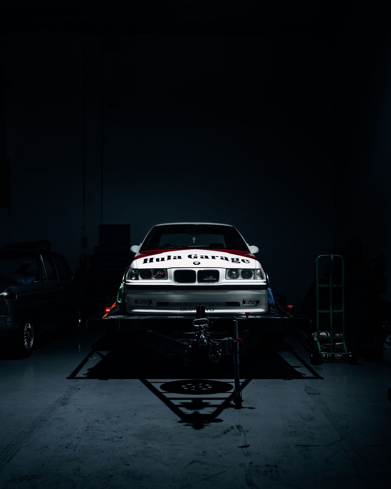 BMW race car with Hula Garage livery on a lift in a dark automotive garage.