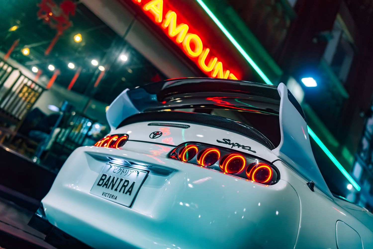 White Toyota Supra with glowing red taillights and custom spoiler parked outside a neon-lit venue at night.
