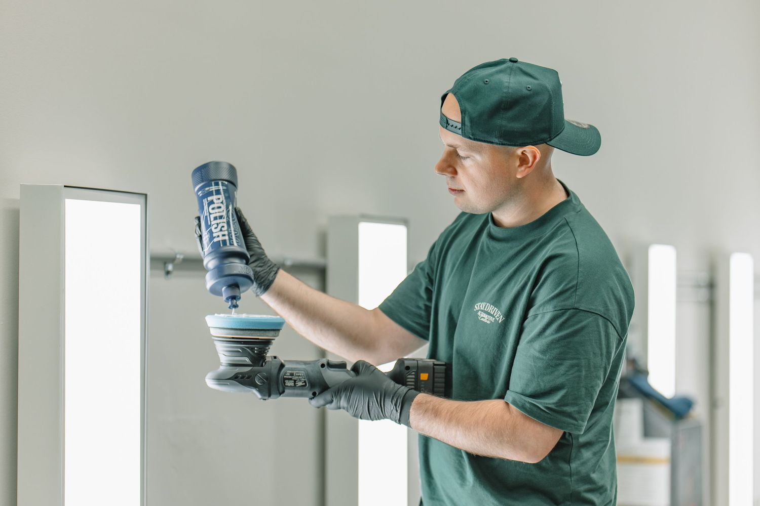A technician in a green cap and shirt uses a Rupes polishing tool on a surface in a professional detailing shop.