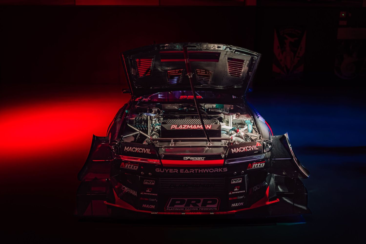 A high-performance drift car with an exposed engine bay, illuminated by dramatic red and blue lighting in a dark studio setting.