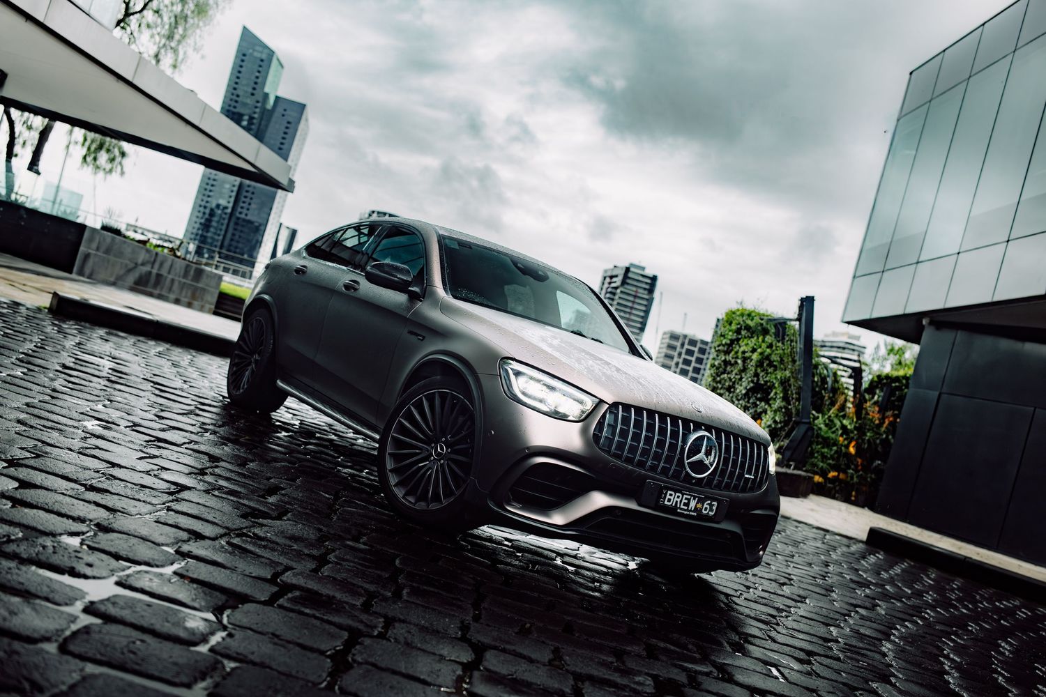 Matte gray Mercedes-AMG GLC SUV parked on cobblestone street with modern glass buildings in background on overcast day.